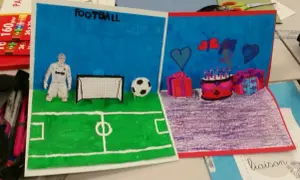 Un dessin pop up de football avec un joueur, un ballon et un but. L'autre partie montre un gâteau d'anniversaire, des cadeaux et des cœurs.