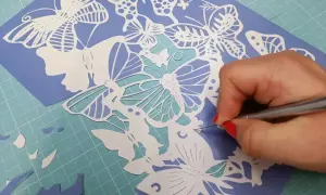 Main d'une personne aux ongles rouges découpant avec précision un motif de papillons en papier. Un tuto de papier découpé en cours.