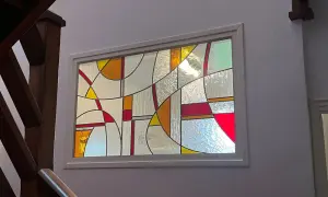 Un vitrail aux formes géométriques abstraites, aux couleurs vives et translucides, éclaire un escalier en bois. Un dessin vitrail facile à admirer.