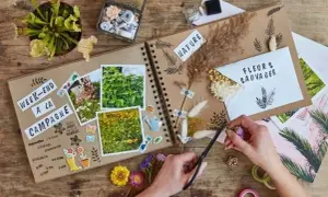 Un carnet de voyages en cours de création, orné de photos de nature, de fleurs séchées et d'autocollants. Une main coupe des brins d'herbe pour décorer.