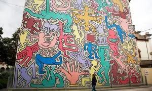 Un mur immense, un peintre bonhomme couleur, des silhouettes vives dansent sur fond blanc. Une femme passe devant cette fresque joyeuse.
