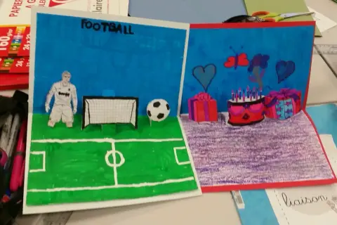 Un dessin pop up de football avec un joueur, un ballon et un but. L'autre partie montre un gâteau d'anniversaire, des cadeaux et des cœurs.