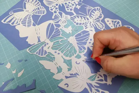 Main d'une personne aux ongles rouges découpant avec précision un motif de papillons en papier. Un tuto de papier découpé en cours.