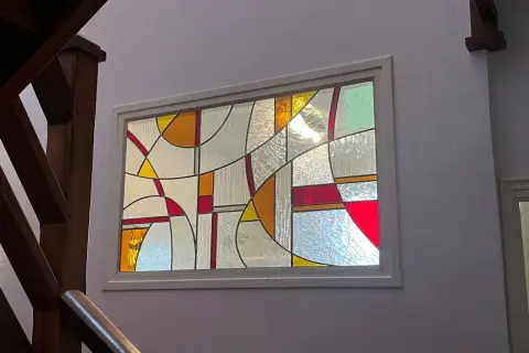 Un vitrail aux formes géométriques abstraites, aux couleurs vives et translucides, éclaire un escalier en bois. Un dessin vitrail facile à admirer.