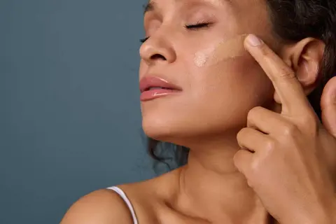 Une femme applique du fond de teint sur sa joue. La **peinture sur la peau** est une étape clé pour un teint parfait.