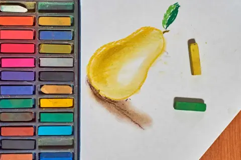 Un dessin au pastel d'une poire jaune avec une feuille verte. Des pastels de différentes couleurs sont disposés à côté.