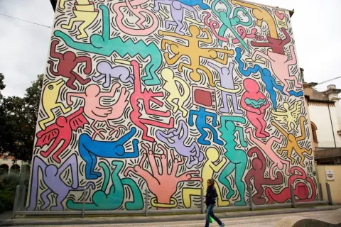 Un mur immense, un peintre bonhomme couleur, des silhouettes vives dansent sur fond blanc. Une femme passe devant cette fresque joyeuse.
