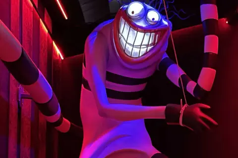 Personnage étrange au sourire démesuré, rappelant un dessin Tim Burton facile. Rayures noires et blanches, lumières rouges et bleues.
