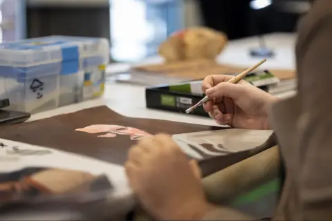 Une main tient un pinceau, peignant un visage sur du papier brun. La scène illustre comment devenir artiste, avec des fournitures d'art en arrière-plan.