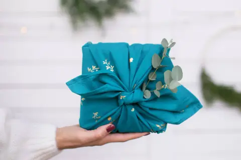 Préparer Noël : un cadeau joliment emballé dans un tissu bleu canard orné de motifs dorés, décoré d'une branche d'eucalyptus.