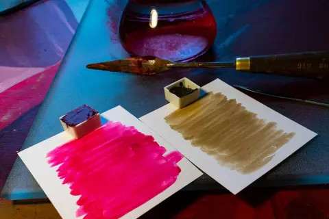 Des petits pots de peinture acrylique, un pourpre intense et un brun terreux, prêts à mélanger pigments. Une spatule et un flacon de liquide rouge complètent la scène.