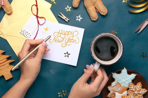 Mains écrivant "Happy New Year" sur une carte, avec une tasse de café et des biscuits.