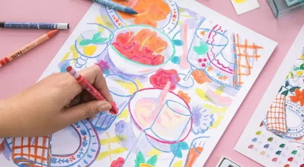 Une main colorie une scène de repas avec des pastels. Une idée art créatif pour un moment joyeux et coloré.