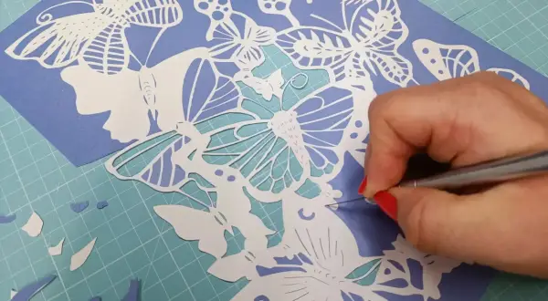 Main d'une personne aux ongles rouges découpant avec précision un motif de papillons en papier. Un tuto de papier découpé en cours.