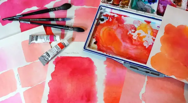 Des carrés de feutre aquarelle aux teintes roses et orangées, des tubes de peinture et des pinceaux. L'utilisation du feutre aquarelle est mise en avant.
