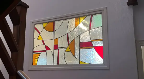 Un vitrail aux formes géométriques abstraites, aux couleurs vives et translucides, éclaire un escalier en bois. Un dessin vitrail facile à admirer.