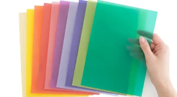 Une main tient une feuille verte translucide, révélant une pile de papiers colorés. Idéal pour faire un vitrail en papier calque.