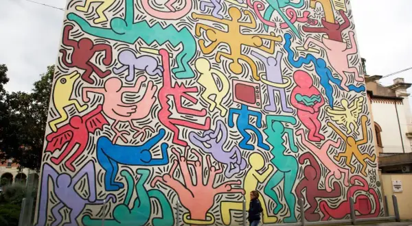 Un mur immense, un peintre bonhomme couleur, des silhouettes vives dansent sur fond blanc. Une femme passe devant cette fresque joyeuse.