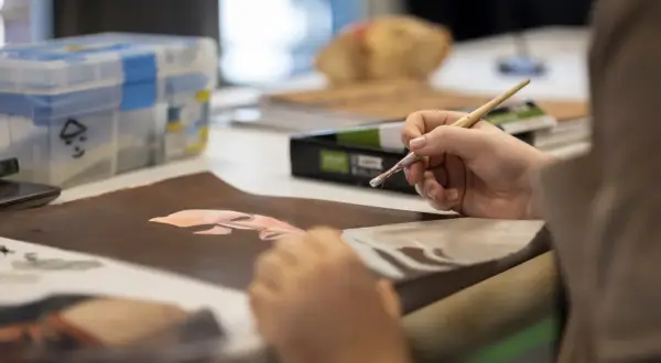 Une main tient un pinceau, peignant un visage sur du papier brun. La scène illustre comment devenir artiste, avec des fournitures d'art en arrière-plan.