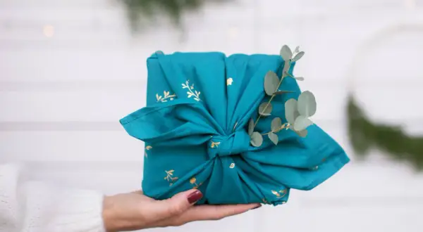 Préparer Noël : un cadeau joliment emballé dans un tissu bleu canard orné de motifs dorés, décoré d'une branche d'eucalyptus.