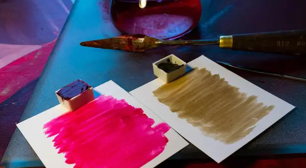 Des petits pots de peinture acrylique, un pourpre intense et un brun terreux, prêts à mélanger pigments. Une spatule et un flacon de liquide rouge complètent la scène.