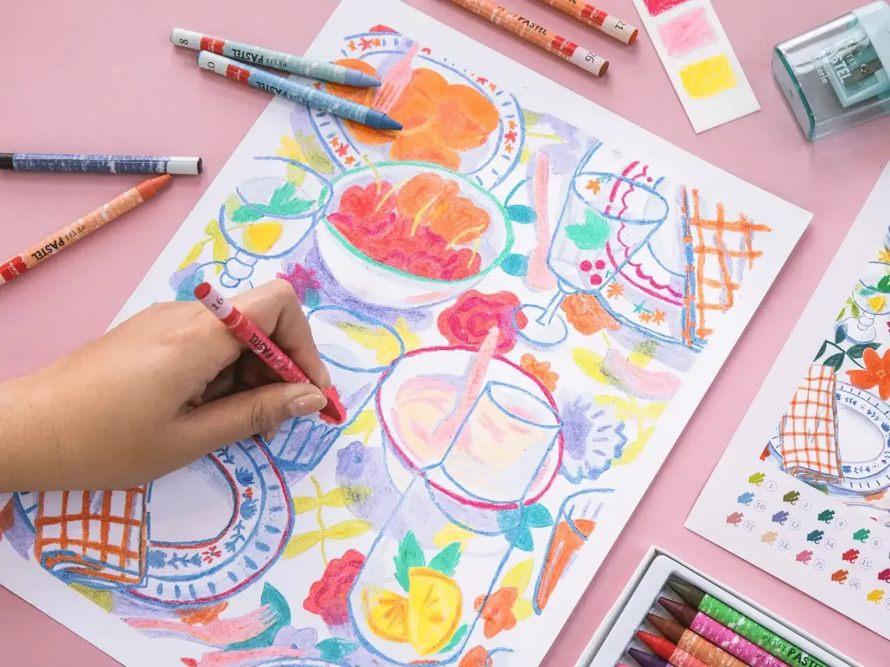 Une main colorie une scène de repas avec des pastels. Une idée art créatif pour un moment joyeux et coloré.