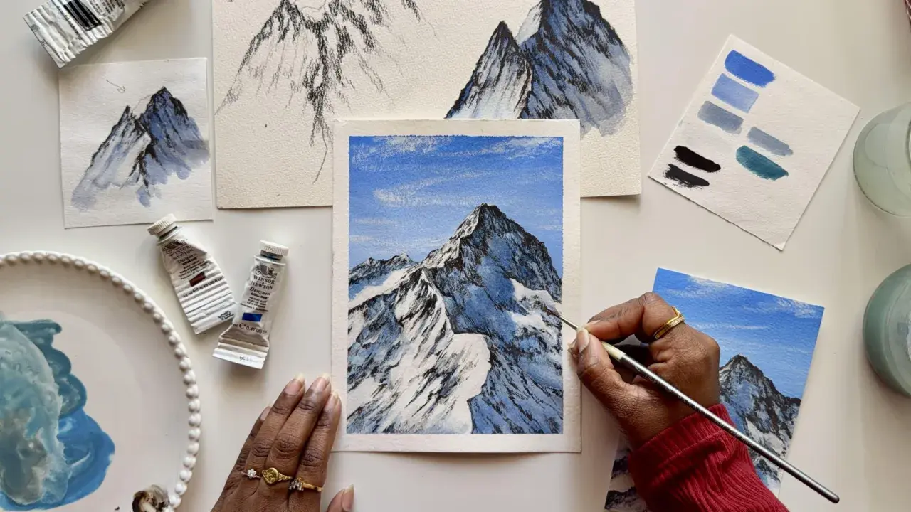 Apprendre comment faire du blanc avec de la gouache pour peindre des montagnes enneigées. Des tubes de peinture, des études et une œuvre en cours sont visibles.
