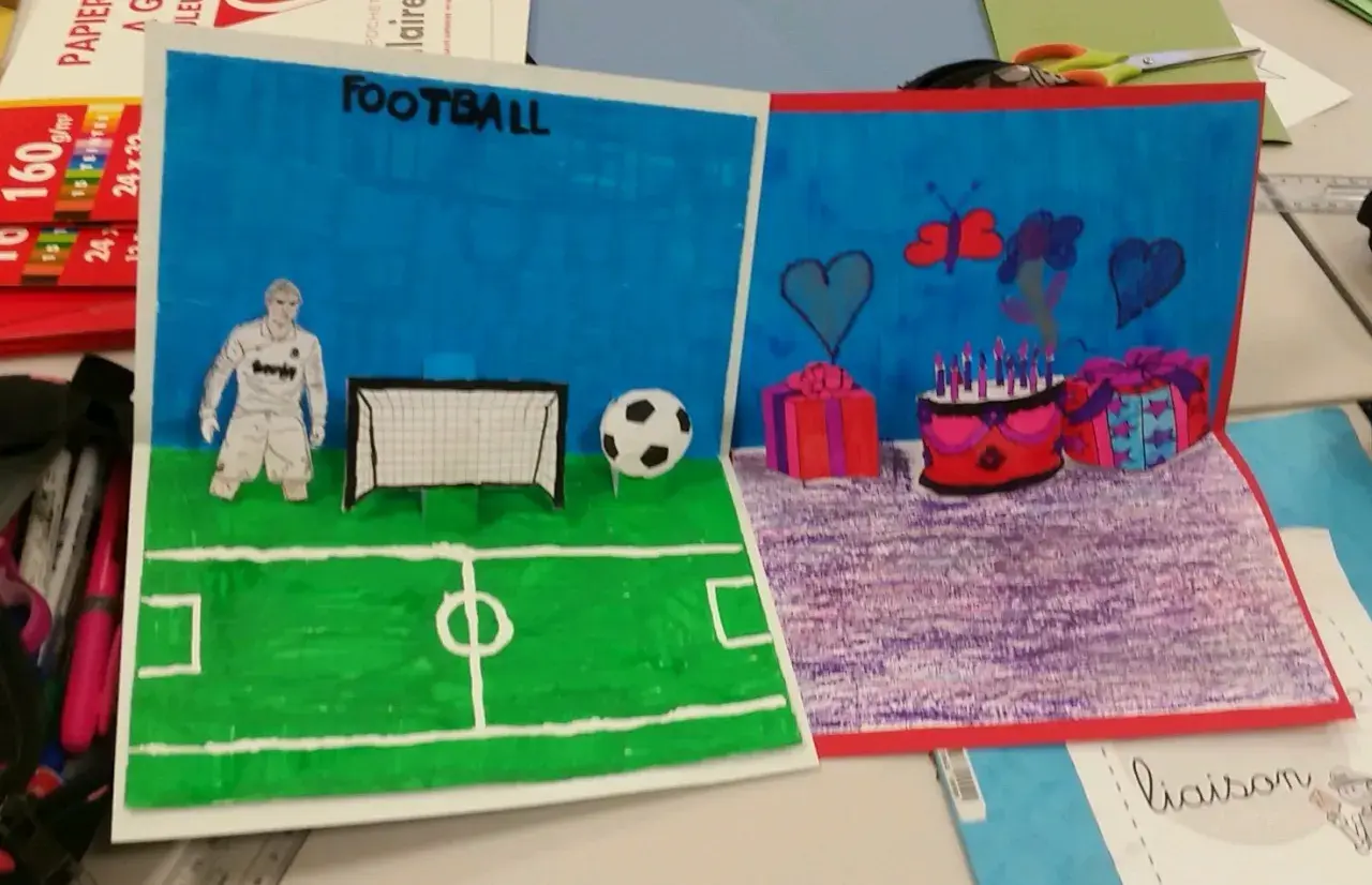 Un dessin pop up de football avec un joueur, un ballon et un but. L'autre partie montre un gâteau d'anniversaire, des cadeaux et des cœurs.