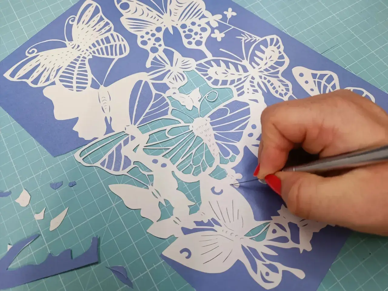 Main d'une personne aux ongles rouges découpant avec précision un motif de papillons en papier. Un tuto de papier découpé en cours.