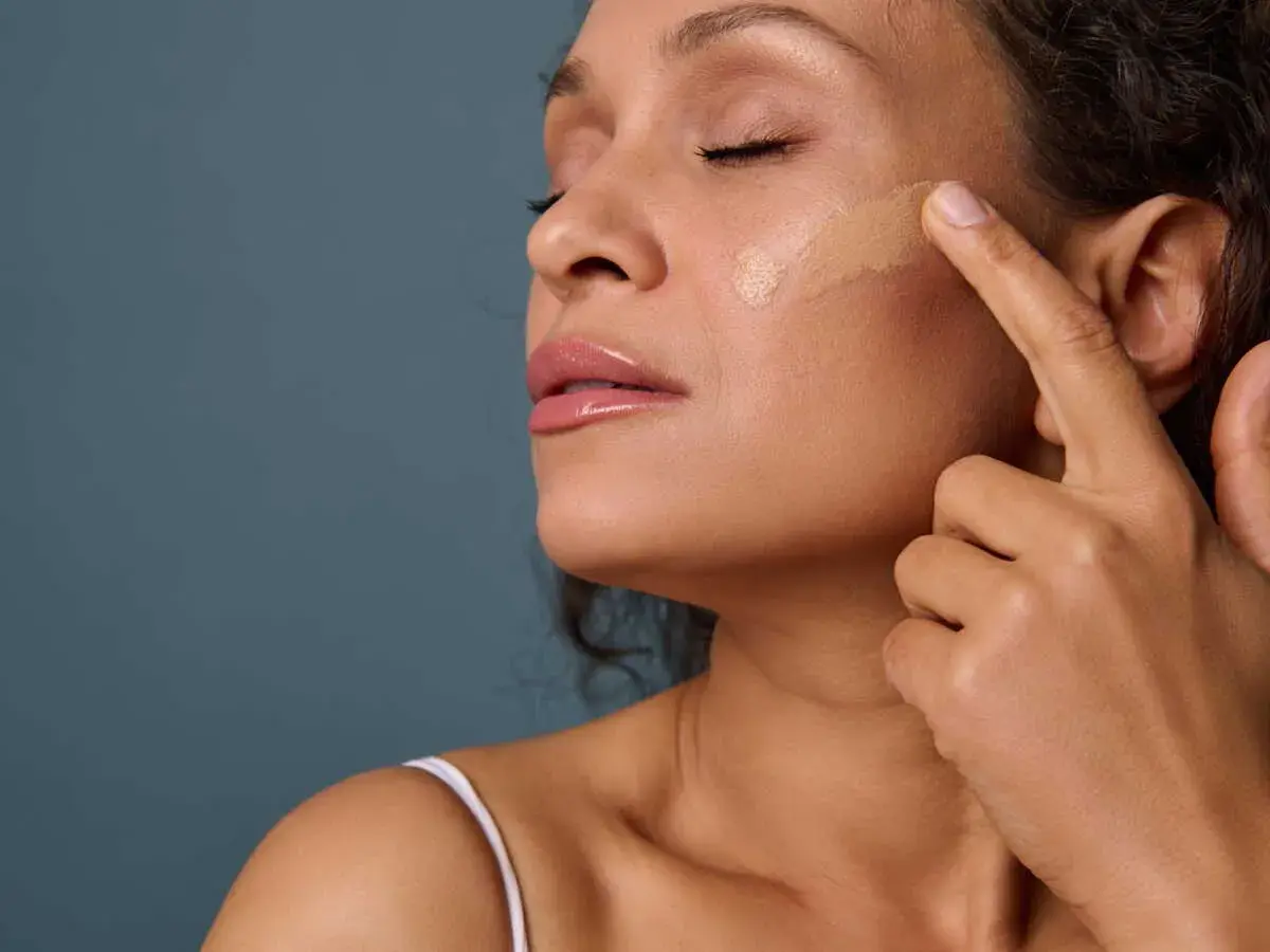 Une femme applique du fond de teint sur sa joue. La **peinture sur la peau** est une étape clé pour un teint parfait.