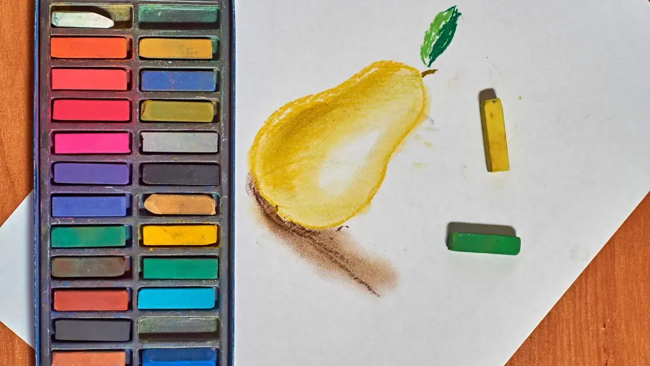 Un dessin au pastel d'une poire jaune avec une feuille verte. Des pastels de différentes couleurs sont disposés à côté.