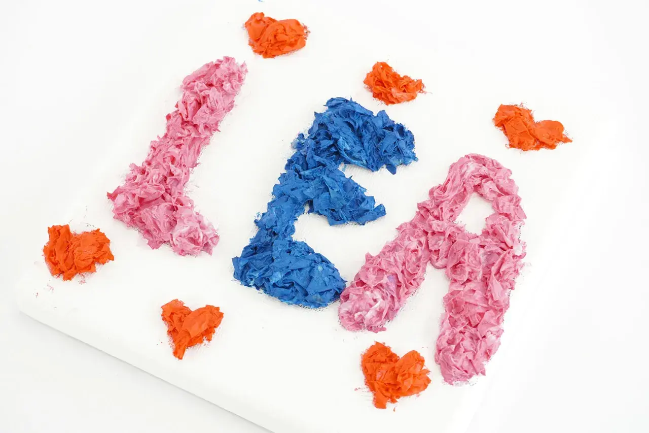 Peinture avec les mains : les lettres "LÉA" en relief, rose et bleu, entourées de cœurs orange sur une toile blanche.
