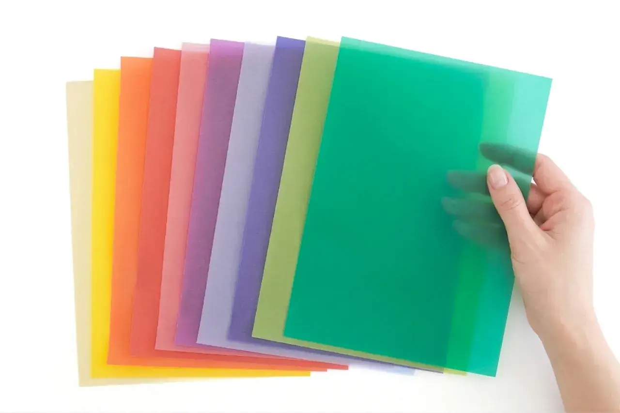 Une main tient une feuille verte translucide, révélant une pile de papiers colorés. Idéal pour faire un vitrail en papier calque.