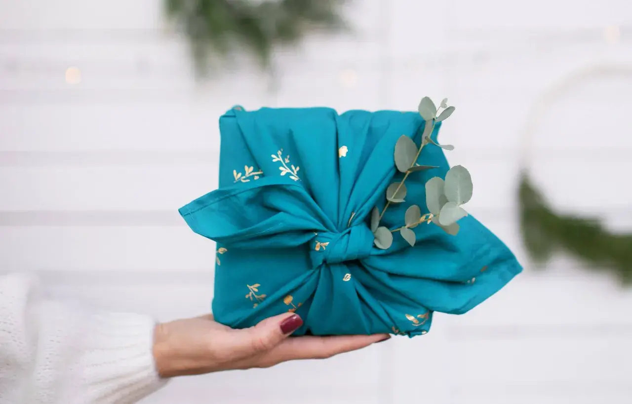 Préparer Noël : un cadeau joliment emballé dans un tissu bleu canard orné de motifs dorés, décoré d'une branche d'eucalyptus.