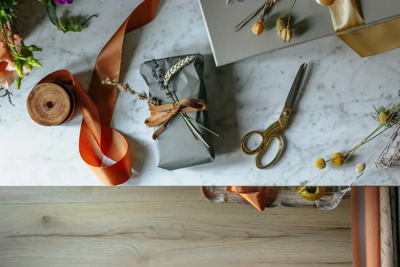 Préparation d'un cadeau pour la fête des grands-mères : un paquet gris décoré de lavande et de blé, entouré de ruban orange et de ciseaux dorés.