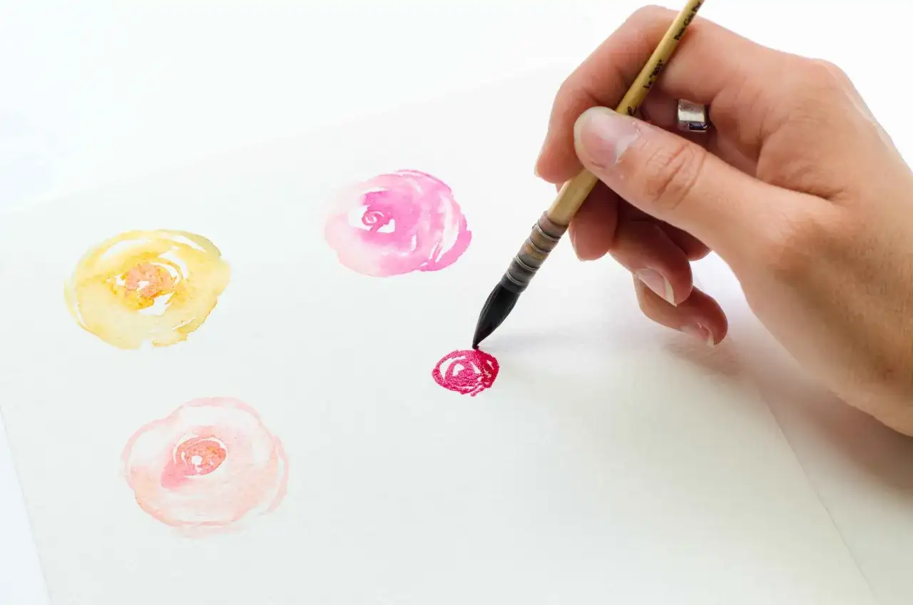 Un dessin aquarelle de fleurs délicates en cours de création. Une main tient un pinceau pour ajouter une touche de rouge vif.