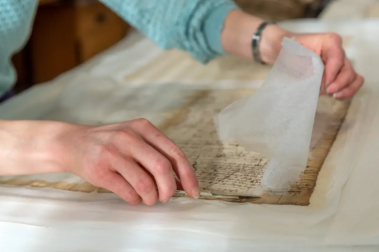 D&eacute;tail d'une restauration : une main d&eacute;licate utilise un scalpel pour comment nettoyer du papier jauni, soulevant une fine feuille protectrice.