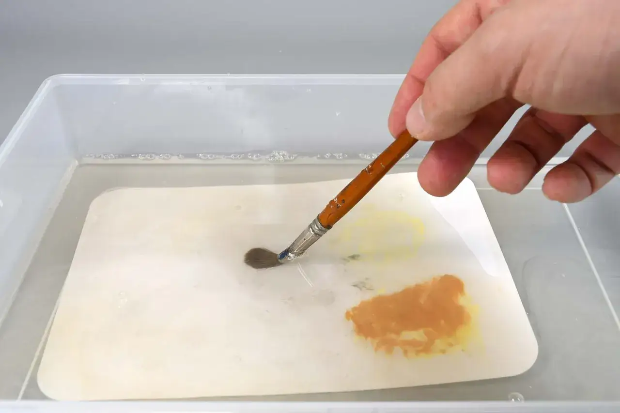 Une main trempe un pinceau dans de l'eau, diluant une peinture fonc&eacute;e qui s'&eacute;claircit en s&eacute;chant sur une palette.