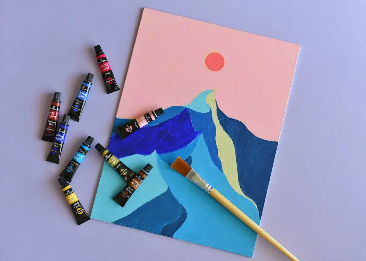 Une id&eacute;e de dessin sur toile facile : un paysage abstrait avec des montagnes bleues et un soleil rose, entour&eacute; de tubes de peinture acrylique.