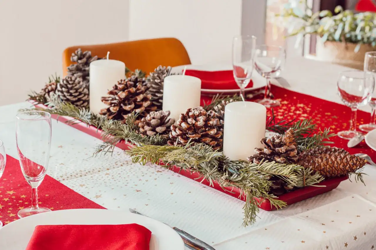 Un chemin de table Noël à faire soi-même orné de pommes de pin, de branches de sapin et de bougies blanches sur une table dressée.