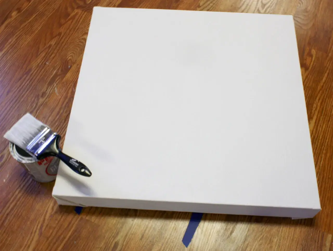 Un pinceau tremp&eacute; dans une bo&icirc;te de peinture est pr&ecirc;t &agrave; &ecirc;tre utilis&eacute; sur un grand support de peinture acrylique blanc, pos&eacute; sur un parquet en bois.
