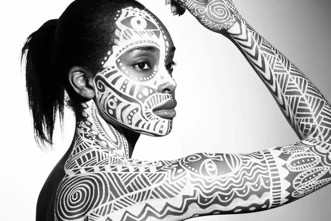 Une femme au visage et au corps couverts de motifs complexes en peinture sur la peau. L'art corporel blanc contraste avec sa peau sombre.