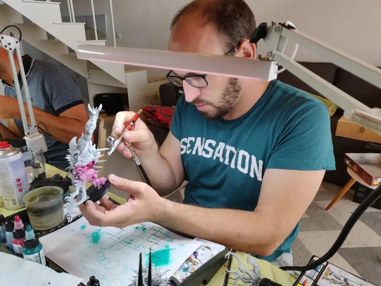 Un homme concentr&eacute; apprend &agrave; peindre une figurine avec un a&eacute;rographe. Des pots de peinture et des pinceaux sont dispos&eacute;s autour de lui.