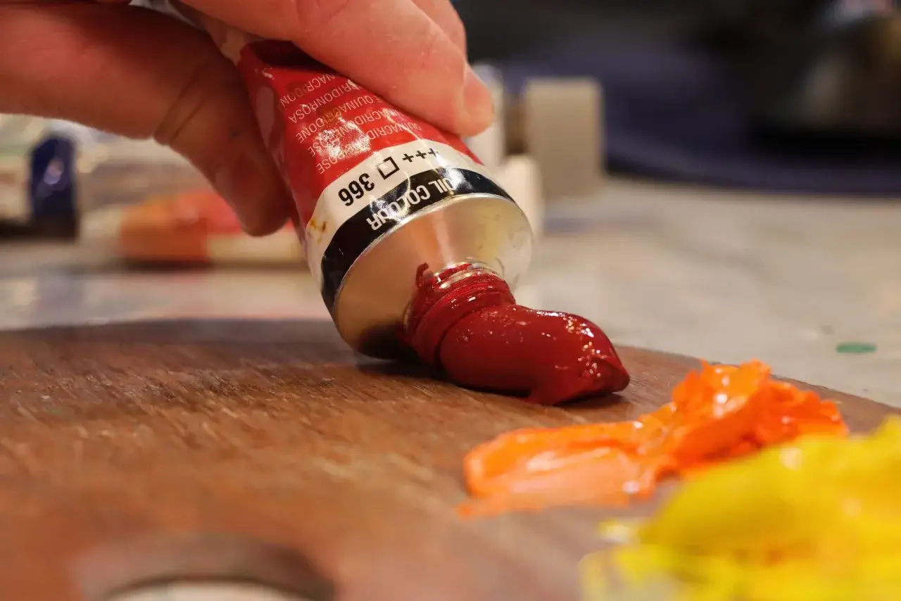 Main rouge sort d'un tube pour se m&eacute;langer aux pigments peinture acrylique sur une palette en bois.