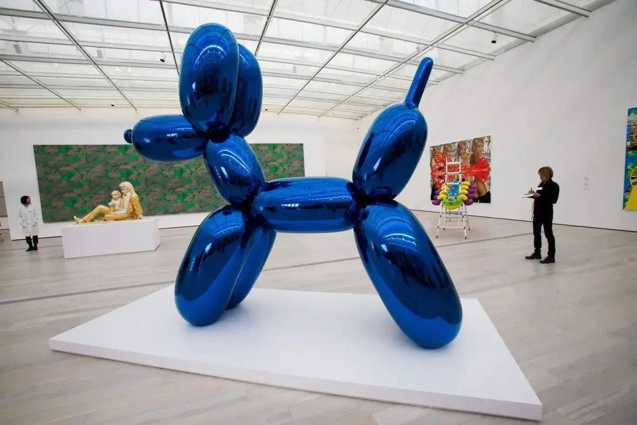 Une &oelig;uvre d'art contemporaine, un chien ballon bleu m&eacute;tallique, tr&ocirc;ne dans une galerie. D'autres &oelig;uvres et visiteurs compl&egrave;tent la sc&egrave;ne.