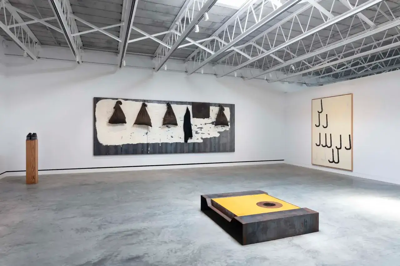 Exposition d'un artiste italien contemporain : une &oelig;uvre murale abstraite, une sculpture m&eacute;tallique jaune et une autre sur un socle en bois.