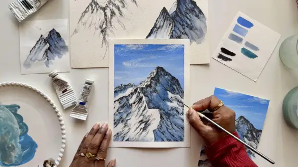 Apprendre comment faire du blanc avec de la gouache pour peindre des montagnes enneigées. Des tubes de peinture, des études et une œuvre en cours sont visibles.