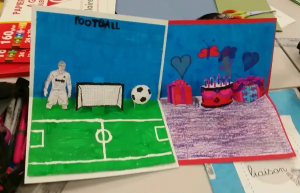 Un dessin pop up de football avec un joueur, un ballon et un but. L'autre partie montre un gâteau d'anniversaire, des cadeaux et des cœurs.