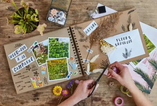 Un carnet de voyages en cours de création, orné de photos de nature, de fleurs séchées et d'autocollants. Une main coupe des brins d'herbe pour décorer.