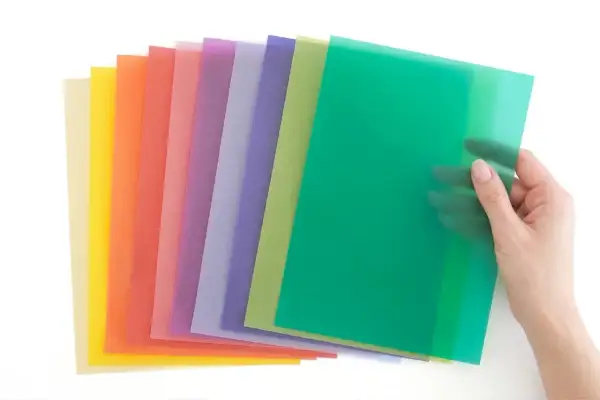 Une main tient une feuille verte translucide, révélant une pile de papiers colorés. Idéal pour faire un vitrail en papier calque.
